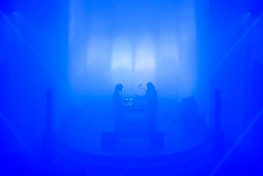 In a completely blue room two silhouettes are standing on a scene